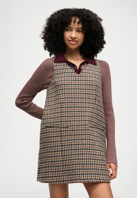 Vestido de padrão pied-de-poule, apresentando um padrão em castanho, preto e creme, com dois bolsos. Combinado com uma camisa de manga longa em burgundy por baixo.