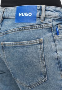 Svetlomodré riflové nohavice s veľkou modrou koženou náplasťou označenou nápisom "HUGO" na páse a kontrastným žltým prešívaním pozdĺž švov.