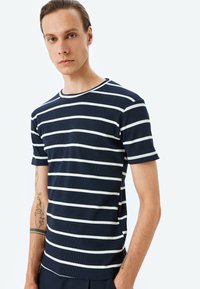 Marineblaue Kurzarm-T-Shirt mit weißen horizontalen Streifen, gerippter Textur, Rundhalsausschnitt und taillierter Passform. Zeigt sichtbare Tattoos am Arm.
