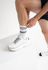 Weiße Sportsocken mit schwarzen diagonalen Streifen und grauen Akzenten werden über einem weißen Sneaker mit strukturierten Mustern adjustiert.