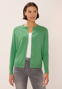 Zelený cardigan s kulatým výstřihem, dlouhými rukávy a zapínáním na knoflíky, vyrobený z měkké látky, doplněný o bílou košili a šedé džíny.