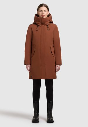 Parka marrón con capucha, cremallera frontal, dos bolsillos laterales y cordones ajustables. Lleva puesta sobre leggings negros y botas de cuero.