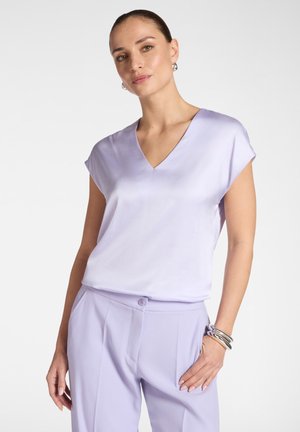 Kobieta ubrana w jasnofioletową bluzkę z krótkim rękawem i dekoltem w serek oraz pasujące wysokie spodnie, do tego srebrne kolczyki koła i bransoletki.