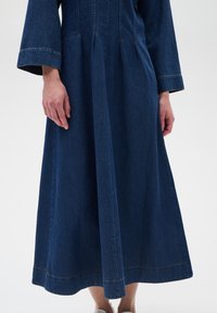 Denim haljina s dugim rukavima, dugog kroja, tamnoplave boje, vidljivim šavovima i blago raširenim suknjom.
