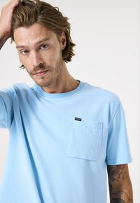 Lichtblauw katoenen t-shirt met korte mouwen, voorzien van een vierkante zak op de linkerborst met een zwart logo-label. Zachte textuur.