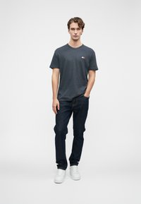 Mladý muž s vlnitými vlasy, oblečený v tmavě šedém tričku, tmavě modrých džínách a bílých teniskách, stojící s jednou rukou v kapse.