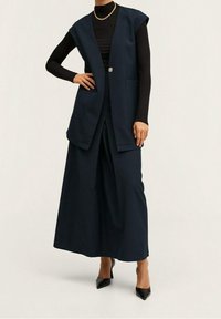 Blazer senza maniche blu navy con un solo bottone, abbinato a pantaloni a gamba larga. Tessuto testurizzato, due tasche frontali e tacchi neri a punta.