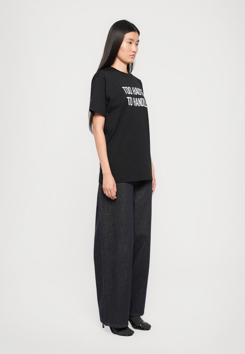 Schwarzes T-Shirt mit weißem Text, der "ZU HEISS ZUM HANDHABEN" liest, kombiniert mit lockeren, dunklen Jeans und schwarzen Schuhen. Glattes Material und lässige Passform.