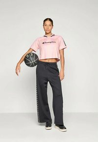 Svetlo roza skrajšana majica s črnim logotipom "Champion", v kombinaciji s črnimi trenirkami z belimi stranskimi akcenti. Model drži košarko.