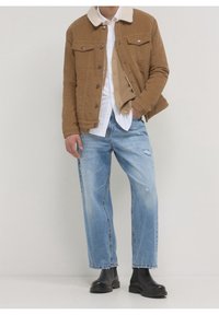 Bruine corduroy jas met een schapenvachtkraag, wit overhemd, beige trui, lichtblauwe jeans met slijtageplekken en zwarte enkellaarsjes.