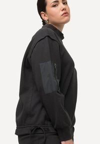 Felpa nera con tessuto testurizzato, presenta una tasca con zip sulla manica sinistra e un orlo regolabile con cordino per una vestibilità personalizzabile.
