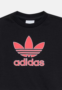 Melnā džemperī ar izteiktu sarkano Adidas logotipu un trefoil dizainu, ar apaļu kakla izgriezumu un teksturētu audumu.