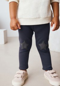 Leggings azul marino con parches de estrellas negras en las rodillas, combinados con un suéter crema con motas y zapatillas de velcro rosa claro.