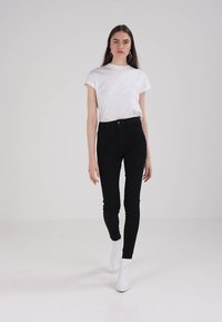 Jeans skinny de tiro alto negros combinados con una camiseta ajustada blanca y botas blancas hasta el tobillo, sobre un fondo blanco liso.