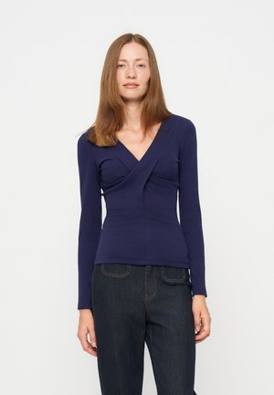 Femme aux cheveux châtain clair et raides portant un haut moulant bleu foncé à manches longues et un jean foncé, debout devant un fond clair uni.
