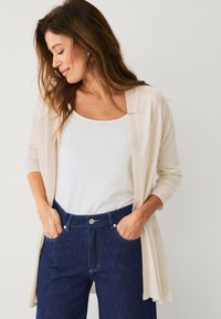 Cardigan beige leggero con maniche lunghe, design davanti aperto; abbinato a una canotta bianca e a jeans in denim blu scuro.