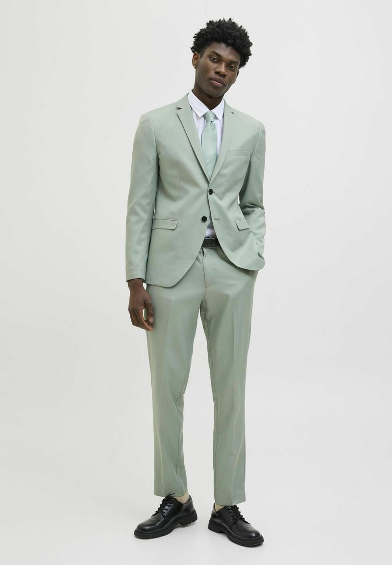 Abito verde chiaro con rever a punta, chiusura a un bottone e tasche laterali. Abbinato a una camicia bianca e a una cravatta coordinata, completato da scarpe nere.