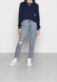 Pull marin à côtes avec col à demi-fermeture éclair, associé à un jean bleu clair déchiré avec des déchirures, et des baskets blanches montantes.