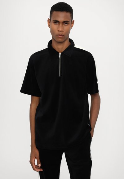 Polo-shirt noir à manches courtes avec col zippé, présentant une texture lisse et un détail en bande blanche sur les épaules.