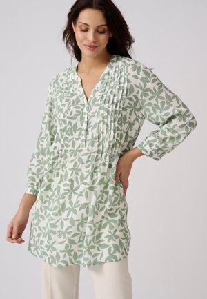Mujer que lleva una blusa túnica blanca con estampados de hojas verdes y detalle plisado en la parte delantera, combinada con pantalones de color claro, posando con una mano en la cadera.