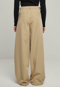 Pantalon large beige avec une taille élastique, deux poches arrière et une texture lisse, légèrement brillante. Convient pour une tenue décontractée.