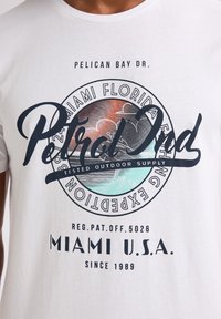 Camiseta blanca de algodón con un gráfico circular que presenta una escena de la playa de Miami, letras en negrita en negro y colores pastel, y elementos de diseño curvados.