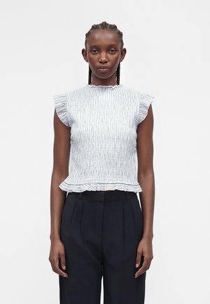 Kobieta z zaplecionymi włosami, ubrana w jasnoszary w paski bezrękawnik z falbankami oraz czarne plisowane spodnie z wysokim stanem, stoi na białym tle.