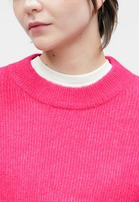 Cilvēks, kuram mugurā ir spilgti rozā ribota džemperis virs baltas kreklu ar apaļu kakla izgriezumu, redzams tikai apakšējais sejas un kakla daļa.