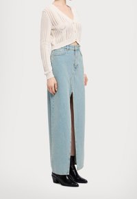 Biały sweter rozpinany na guziki z dekoltem w serek, ozdobiony teksturowanymi detalami. Jasnoniebieska spódnica z denimu z przednim rozcięciem, zestawiona z czarnymi botkami na obcasie.