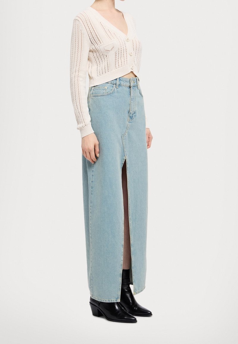 Biały sweter rozpinany na guziki z dekoltem w serek, ozdobiony teksturowanymi detalami. Jasnoniebieska spódnica z denimu z przednim rozcięciem, zestawiona z czarnymi botkami na obcasie.