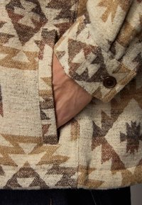 Veste en mélange de laine avec un fond crème et des motifs géométriques bruns. Poignet boutonné avec une poche ; tissu texturé.