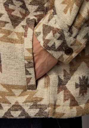Wool blend jacket featuring a cream background with brown geometric patterns. Buttoned cuff with a pocket; textured fabric.
