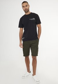 T-shirt nera a maniche corte con testo bianco, abbinata a pantaloni cargo verdi oliva. Il modello indossa sneakers bianche. Tessuto morbido e vestibilità comoda.