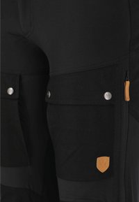 Pantalons cargo noirs avec deux poches latérales, boutons-pression argentés et tirette de fermeture éclair orange contrastante. Présente un tissu texturé et un patch logo.