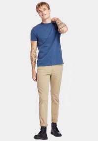 Tmavě modré tričko s krátkým rukávem, béžové úzké kalhoty, černé kožené boty se šněrováním, stojící póza, světlé pozadí, ukazující tetování na pažích.