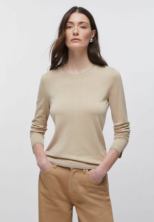 Frau mit langen braunen Haaren, die einen beigen Pullover mit langen Ärmeln und beige Hose trägt, Hände in den Taschen, stehend vor einem schlichten hellen Hintergrund.