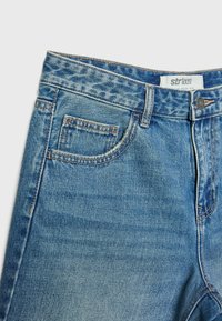 Jean en denim bleu en gros plan montrant la poche avant, la ceinture avec passants, le bouton et l’étiquette de marque cousue à l’intérieur.