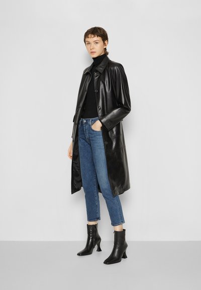 Schwarzer Lederrock mit durchgeknöpftem Vorderteil, langen Ärmeln und gerader Passform, kombiniert mit blauen Jeans und schwarzen Ankle Boots mit Absatz.
