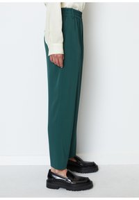 Tealfarbene, elastische Hosen mit geradem Schnitt und glatter Textur, kombiniert mit glänzenden schwarzen Slippern mit dicker Sohle.