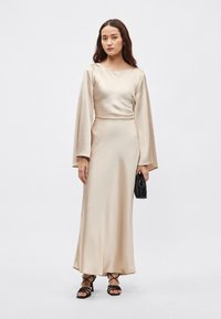 Šviesiai smėlio spalvos satino ilga suknelė su ilgomis rankovėmis, aptempta liemeniu, laisvo silueto. Modelis laiko mažą juodą rankinę, derina su juodais bateliais su dirželiais.