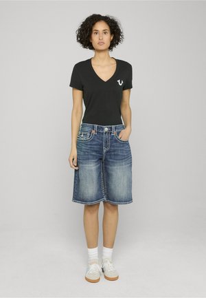 Schwarzes V-Ausschnitt-T-Shirt mit kleinem Logo, kombiniert mit einem knieumspielenden blauen Jeansrock mit dekorativen Nähten und Fronttaschen.