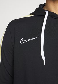 Czarna bluza z kapturem z białym sznurkiem i białym logo Nike na klatce piersiowej, z białymi i jasnoskalnymi paskami na ramieniu.