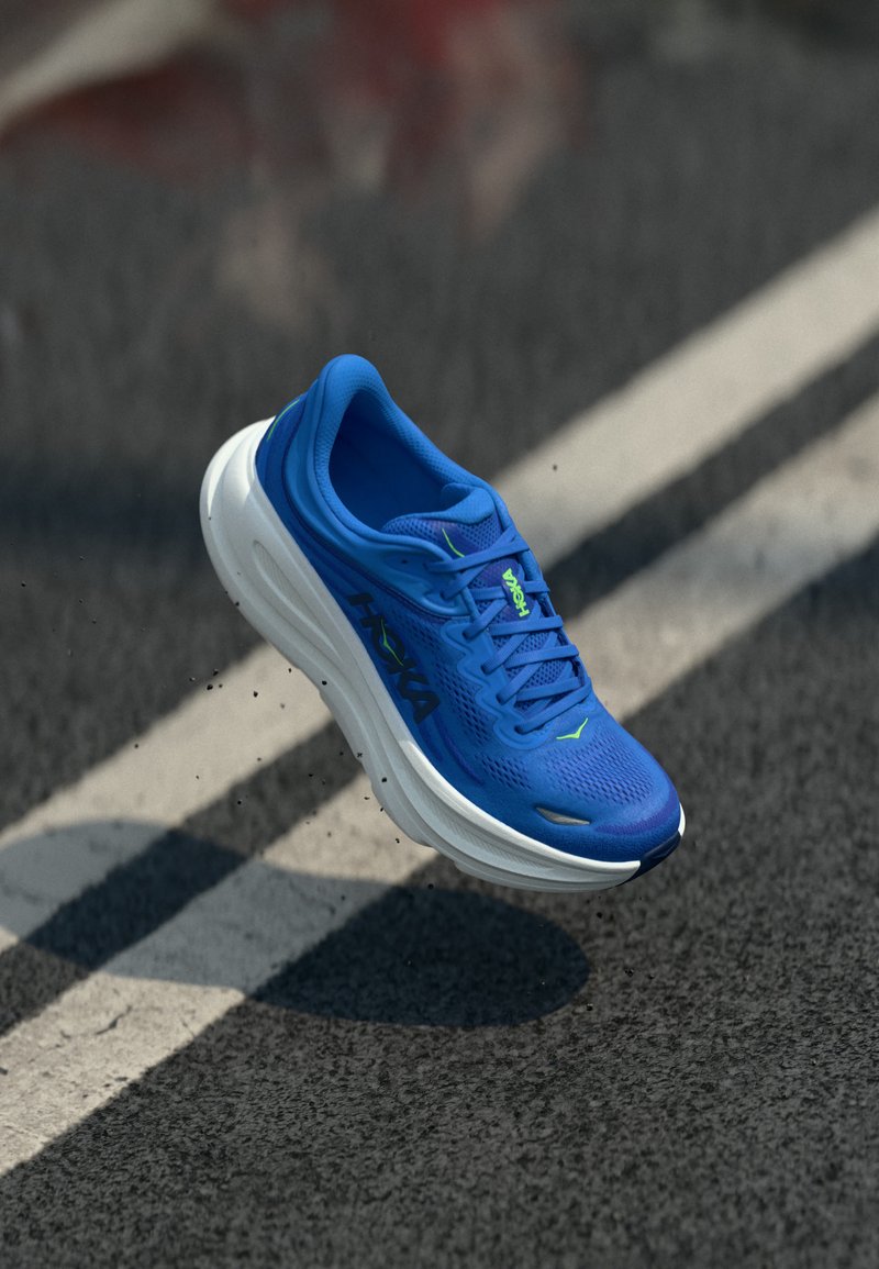 Μπλε αθλητικό παπούτσι με λευκή σόλα στον αέρα πάνω από ασφάλτινο δρόμο με λευκές γραμμές, που δείχνει το όνομα της μάρκας στο πλάι και στη γλώσσα.