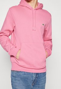 Hoodie in cotone rosa con tasca a canguro, lacci e un piccolo logo sul petto. Il tessuto appare morbido e texture.