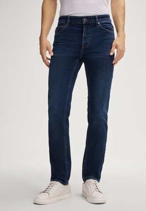 Dark blue denim jeans with a straight-leg fit, five-pocket design, and a button fly, paired with white sneakers on a neutral background.