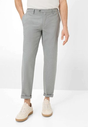 Homme portant un pantalon slim gris clair retroussé aux chevilles et des baskets blanc cassé, debout avec une main dans la poche sur fond blanc.