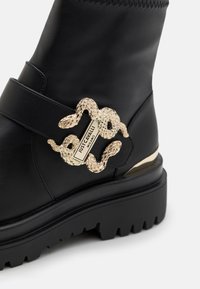 Botín negro de cuero hasta el tobillo con suela gruesa y una hebilla en forma de serpiente dorada con un detalle de logo grabado. Parte superior texturizada con acento de correa.