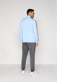 Casaco desportivo azul-claro com fecho de correr e gola alta, com riscas cinzentas nas laterais, combinado com calças de fato-de-treino cinzentas e sapatilhas cinza-claro.