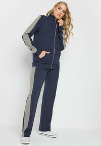 Tuta navy con giacca con zip e pannelli grigi sulle spalle; pantaloni abbinati con strisce grigie sui lati; realizzata in tessuto morbido e confortevole.