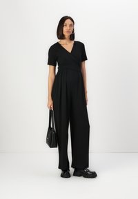 Combinaison noire avec un devant drapé, des manches courtes et un design à jambes larges. Assortie avec des bottes noires chunky et un petit sac à main noir.